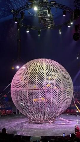 A slow, static shot captures a circus performance inside a large, geodesic sphere. The sphere is a metallic cage with diamond-shaped openings, casting a pinkish glow from unseen lights. Inside, several motorcyclists are visible riding in a circular motion, their bikes appearing as yellow blurs against the cage. The sphere is situated on a circular stage, lit with purple and white spotlights. In the background, rows of red stadium seats are visible, with a large audience seated and looking towards the performance. The ceiling of the circus tent is dark blue and adorned with a circular lighting rig that hangs above the stage. The overall atmosphere suggests a thrilling and dramatic circus act.
