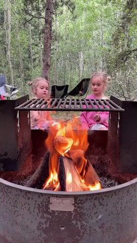 Two young girls are sitting in front of a campfire, roasting marshmallows. The girl on the left is wearing a pink and white plaid shirt and the girl on the right is wearing a pink sweatshirt. They are both holding sticks with marshmallows over the fire. The campfire is in a metal fire pit, and there are trees and foliage in the background. The scene appears to be in a wooded area, possibly a campsite.