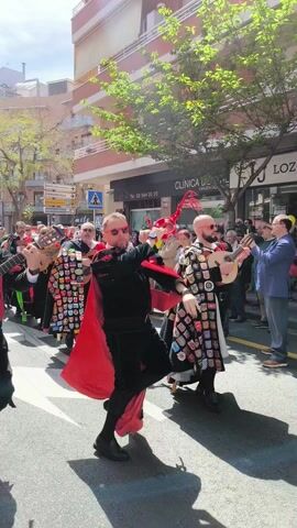 A group of men in costume are marching in a parade down a city street. The men are wearing black capes with colorful patches and red accents. Several of them are playing stringed instruments, including guitars and bandurrias. A man in the front is dancing and twirling a red scarf. The crowd is watching and filming the parade. Buildings and businesses line the street. Trees add greenery to the scene.