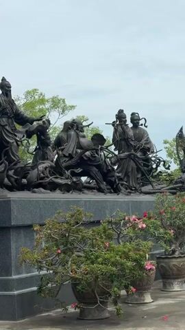The video displays a bronze statue depicting several figures in traditional Chinese attire. In the foreground, a large, ornate bronze lion statue is positioned to the left, facing right. Behind it, a tiered granite pedestal supports the main bronze sculpture. The sculpture features at least five human figures, two prominent ones standing on the left with long beards and elaborate headdresses, seemingly gesturing with their hands. They are surrounded by other figures and what appear to be swirling, organic forms or creatures. To the right of the main figures, a smaller group of figures is visible, one holding a fan and others in discussion. The background consists of lush green trees and a cloudy sky. In the lower right of the frame, several potted plants with pink flowers add a touch of color. The video pans slightly to the right, offering a broader view of the statues and their surroundings.