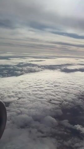 The video presents a view from above a thick layer of clouds, likely taken from an airplane window. The cloud cover is dense and billowy, with varying shades of gray and white, suggesting different cloud densities and depths. Above this sea of clouds, the sky is visible with a mix of light and dark grey cloud formations, some appearing in long, horizontal streaks. The sunlight is subtly diffused, hinting at its presence behind the upper cloud layers.