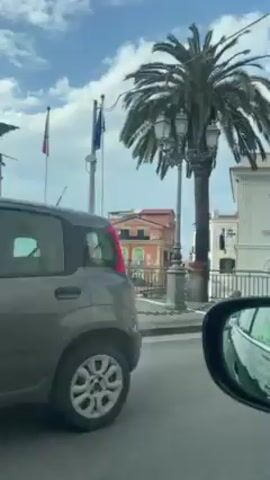 A car drives past a row of buildings, the passenger side window visible in the side view mirror. There is a palm tree near a black metal railing along the side of the road. Two buildings stand to the right, a smaller pink one behind a large white one with a balcony. A gray car drives past on the right. The sky is partly cloudy.