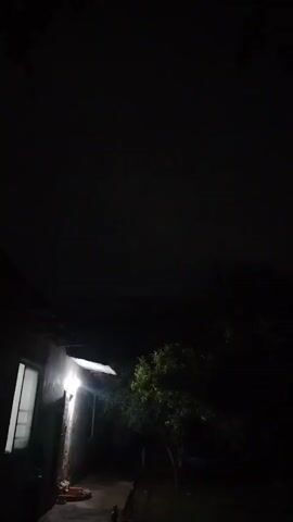 The video shows a dark night scene. A light shines on the side of a building, illuminating a window and a small section of the wall. To the right of the building, a tree is visible, its leaves dark against the night sky. The sky itself is mostly dark, with some faint clouds. The camera pans slightly, showing more of the dark surroundings.