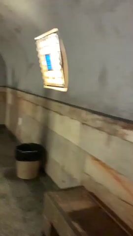 The video shows a group of three people walking down a tiled platform of what appears to be a subway or museum. The platform has an arched ceiling, and windows are intermittently spaced along the wall. The wall itself consists of a textured base and plain wall above. A yellow safety line is painted on the platform's edge. An old blue subway car sits on tracks along the platform. People are walking in the same direction.