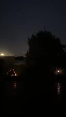 The video captures a dark, ominous scene, likely at night, with a stormy or overcast sky. In the foreground, a wet, reflective surface, possibly asphalt or pavement, suggests recent rain. To the left, a piece of equipment, possibly a wheelbarrow, is tipped over and appears to be emitting sparks or glowing embers, creating a small, bright orange light source. Behind this, a dark tree or dense foliage dominates the center of the frame, silhouetted against a faint light in the upper left corner, which could be a distant streetlamp or building light. To the right, a structure with a dimly lit window or opening casts a faint, yellowish glow. The overall atmosphere is one of eerie stillness and potential danger, enhanced by the darkness and the unusual light sources.