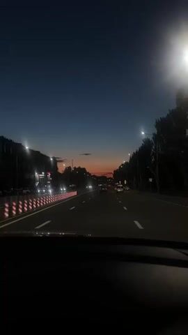 The video shows a view from inside a car driving on a road at dusk. The sky is dark blue with hints of orange and pink on the horizon. Streetlights illuminate the road, and car headlights are visible in the distance. Trees line the right side of the road, and buildings are visible on the left. The car is moving forward, and the perspective suggests a typical road trip or commute. There are no people visible, and the focus is on the scenery and the act of driving.