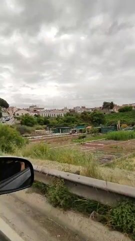 The video captures a panoramic view from a moving vehicle, evidenced by the car's side mirror visible in the lower left of the frame and the passing landscape. The scene unfolds under an overcast sky, filled with heavy grey clouds, suggesting a muted daylight.

In the foreground, a metal guardrail runs horizontally, indicating the vehicle is moving alongside a road. Beyond the guardrail, there's a lush expanse of green vegetation, including tall, wild grasses, bushes, and trees. In the middle ground, several cultivated plots or allotment gardens are visible, some covered with white protective sheets. A distant, faint human figure can be seen near these plots around the 0:02 mark.

Further in the background, a cluster of residential buildings, predominantly light-colored, are nestled on a gentle slope, possibly part of a town or village. More trees dot the landscape beyond the houses, extending towards the horizon. The setting combines elements of a rural and semi-urban environment, consistent with the provided context of Castellar del Vallès, Spain. No discernible text is visible within the frame. The overall atmosphere is tranquil, with the cloudy weather casting a soft light over the landscape.