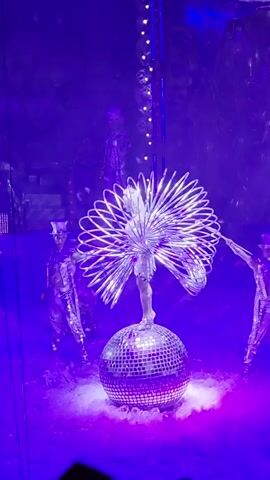 A performer stands balancing on a disco ball, wearing a silver costume with a large, fan-like headdress made of metallic hoops. Several other performers in similar silver costumes and top hats are visible in the background, with one on the left and two on the right. The stage is lit with a strong purple light, and there is a misty or smoky effect around the disco ball. A string of lights hangs from the ceiling in the background. The video appears to be a short clip from a live performance.