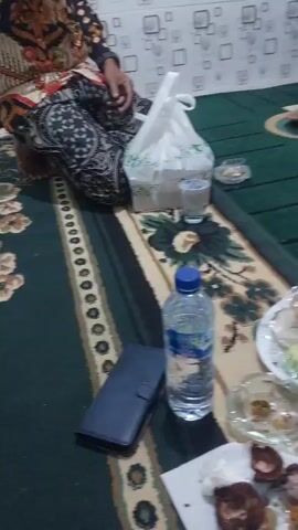 The video shows a group of people seated on a carpeted floor. A white plastic bag with green markings is being held by someone on the left side of the frame, obscuring part of the view. Next to the bag is a glass of water. In the center of the frame is a plastic bottle of water. To the right of the water bottle is a plate of assorted pastries, some green and some yellow. Below the pastries is an ashtray with a few small items in it. In the foreground, there is a white piece of paper with some brown, broken pieces of fruit, possibly mangosteen, and some crumpled napkins. A dark blue wallet is placed next to the water bottle. In the background, a person in a black garment is visible on the right, and another person in a patterned garment is seated on the left. The floor is covered with a carpet that has a dark green and beige striped pattern. A section of the wall behind the people has a tiled pattern with white squares. The lighting appears to be artificial, casting some shadows. The overall atmosphere suggests a casual gathering, possibly a meal or a break.