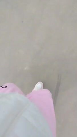 The video shows a person walking on a paved path in a park. The person is wearing white sneakers and pink pants, and the camera is angled down, showing their feet as they walk. The path is surrounded by grass and trees, some of which are bare. There are also lampposts along the path. The sky is overcast, and the wind appears to be blowing.