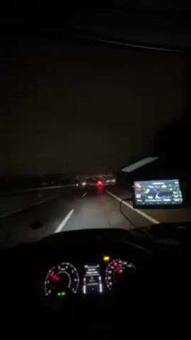 The video shows a first-person view from inside a car driving on a highway at night. The car is moving at a moderate speed, as indicated by the illuminated dashboard. Other vehicles, including cars and a truck, are visible on the road ahead, their headlights and taillights creating bright spots against the dark background. The highway passes under a structure, momentarily obscuring the view of the city skyline in the distance. The cityscape is dotted with lights, adding a contrasting glow to the dark, cloudy sky above. The driver's hands are briefly visible on the steering wheel. The interior of the car is dimly lit, with a navigation screen displaying a map on the dashboard. Overall, the scene depicts a night drive on a highway near a city.