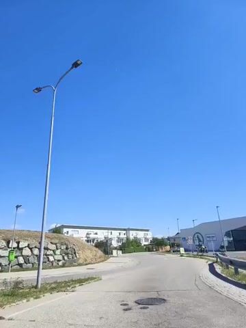 The video shows a street view with a clear blue sky. A modern streetlight with two lamps stands prominently in the foreground. To the left of the streetlight is a rock wall with vegetation on top. A small green bin is visible near the rock wall. A road curves to the right in the middle ground, leading towards buildings in the distance. Another streetlight is visible further down the road. The scene appears to be a suburban or commercial area on a sunny day. There are no people visible in the video.