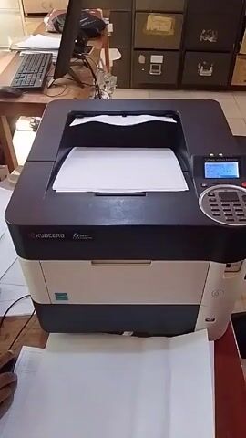 The camera is focused on a Kyocera Ecosys P3045dn printer. It is sitting on a wooden desk. In front of the printer is a hand holding a piece of paper. Behind the printer is a desk with a computer, keyboard, and mouse. Behind that is a set of dark file cabinets. The screen on the printer is lit up.