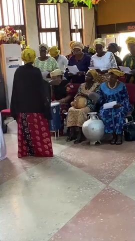 The video shows a group of women, likely a choir, gathered indoors. They are all dressed in vibrant, traditional African attire, predominantly in shades of yellow, blue, green, and red, with matching headscarves. They appear to be singing or performing, with some holding papers, possibly sheet music or lyrics. One woman in the foreground, facing away from the camera, wears a black jacket over a red and black patterned skirt and a yellow headscarf. To her right, another woman is playing a blue drum. Several other women are seated, looking engaged in the activity. In the background, there are windows with decorative grilles, and a sign visible on the wall reads "FAVOUR". The setting appears to be a church or a similar community hall, given the pews and the overall atmosphere. The lighting suggests it is daytime. The mood seems festive and communal.