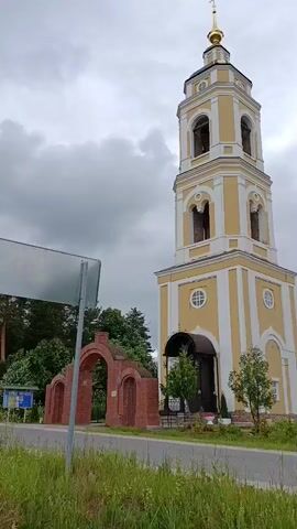 The video shows a church bell tower with a yellow and white exterior, topped with a golden dome and cross. To the left of the bell tower is a partially constructed brick archway. Behind the bell tower and to the right is the main body of the church, also with a golden dome. The sky is overcast, and there is lush green grass and trees in the foreground. The camera pans slightly to the right, revealing more of the church.