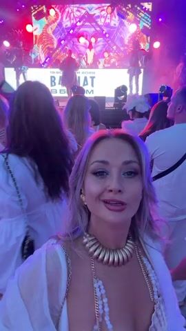 A blonde woman in a seashell-themed bikini top and a gold necklace is in the foreground of the video. She is smiling and gesturing towards the camera while holding a drink. In the background, a crowd of people, mostly wearing white, are gathered around a stage where a band is performing. The stage is lit with vibrant pink and purple lights. The overall atmosphere appears to be a concert or festival.