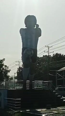 The video shows a statue of Diego Maradona in an Argentina soccer jersey, juggling a soccer ball with his left knee. It is outdoors with overhead power lines and a utility pole in the background. There's also a covered structure with a pickup truck parked beside it in the background. The weather appears to be sunny.