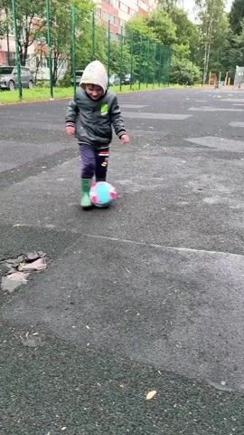 A young child, wearing a hooded jacket, dark pants, and green boots, is playing with a soccer ball on a wet, paved outdoor area. The child kicks the ball repeatedly, sometimes missing it. The background shows a building, trees, and a fence. The weather appears to be overcast or rainy.