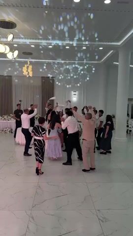 A group of people are dancing in a brightly lit, spacious room with a white marble floor and white walls. The ceiling features recessed lighting and decorative light fixtures. Several people, mostly men dressed in white shirts and dark pants, are raising their arms and waving white cloths. Some women are also dancing, wearing dresses in pastel colors and black and white patterns. The atmosphere appears festive, possibly a wedding reception or a similar celebration. The camera is positioned at a medium distance, capturing the general scene.