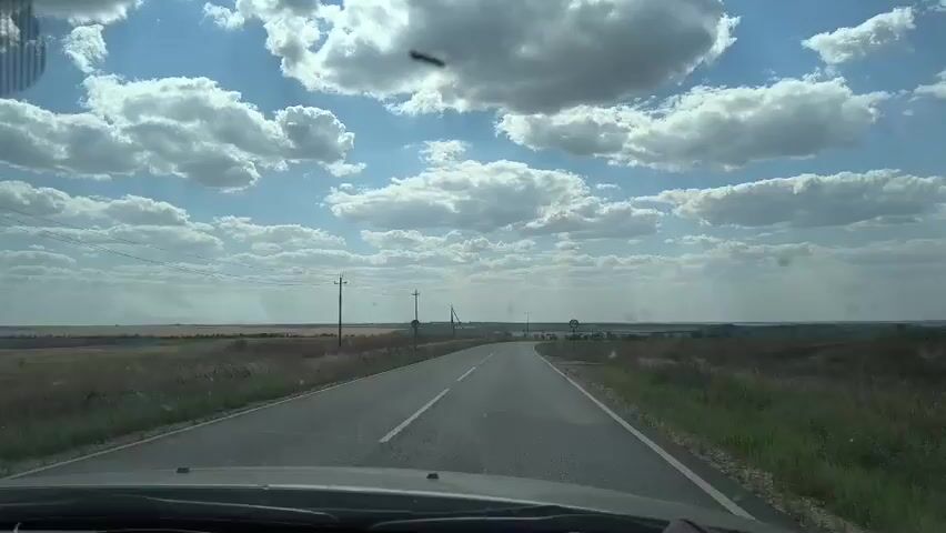 The video shows a view from inside a vehicle traveling on a paved road through a rural landscape under a partly cloudy sky. The road is asphalt with white lane markings. Fields of dry grass and some green vegetation are visible on either side of the road. Utility poles are sparsely placed along the roadside. In the distance, a small settlement or village can be seen. The camera appears to be mounted on the dashboard or windshield of the car. There are no visible occupants of the vehicle. The overall impression is of a journey through a rural, open area on a bright day.