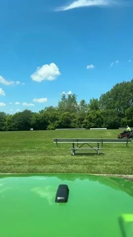 A wide shot captures a bright, sunny day in a grassy park. The sky is a vibrant blue with a few scattered white clouds. In the foreground, the shiny green hood of a vehicle reflects the sky.

The middle ground of the scene features a vast expanse of well-maintained green grass. A few picnic tables with benches are visible, some appearing empty and others with people enjoying the outdoors. In the distance, a person on a riding lawnmower is actively cutting the grass, their bright yellow shirt standing out against the green landscape. Behind the lawnmower, a line of trees forms a dense, green border, suggesting the park is surrounded by woods.

Further back, more trees are visible, with a few glimpses of what might be structures or additional park amenities peeking through. A sign with a blue wheelchair symbol is planted in the grass, indicating a disabled parking or access area.

The overall mood is peaceful and idyllic, suggesting a pleasant day for outdoor recreation. The time of day appears to be midday, given the bright sunlight and the presence of shadows. The location cues, such as the picnic tables, benches, and the open grassy field, point to a public park. The activity taking place is lawn maintenance and enjoying a park setting. There is no visible text other than the wheelchair symbol on the sign.