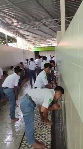 The video shows a group of young men in school uniforms using a series of outdoor faucets to wash their faces. The area appears to be a school or religious institution. The men are all wearing white shirts with green accents and grey pants, suggesting a uniform. They are standing at a long trough-like sink with multiple faucets, and the floor is wet, indicating recent use. The environment is covered by a roof, and the wall behind the sinks is tiled. The overall scene depicts a communal washing ritual, possibly for prayer or hygiene.