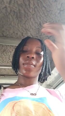 A young Black woman with dark braided hair and a pink t-shirt with a graphic print is in a vehicle. She is looking at the camera, touching her hair and face. The interior of the vehicle is visible in the background.