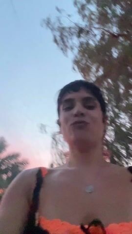 A woman in an orange and black top is filming a video of herself and a man. They are outdoors with trees and a pink and orange sunset in the background. Other people are walking around them. The man is wearing a white shirt and a hat and smiles. The woman excitedly points at the camera.