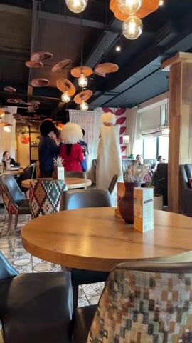 The video pans through a dimly lit, rustic-themed restaurant with multiple tables and chairs. The decor features exposed wood, patterned upholstery on the chairs, and circular, cloud-like light fixtures hanging from the ceiling. On the right side of the screen, a round wooden table is in the foreground. A brown ceramic vase containing dried wheat stalks and a small informational pamphlet are placed on this table. In the background, three individuals stand near a white draped partition. Two of them are wearing large, white, fluffy hats. One person in the center wears a red jacket, and another to their left is dressed in dark clothing. The person to the far right is wearing a cream-colored, long garment. Several other patrons are visible sitting at tables in the background, some of whom appear to be engaged in conversation or dining. The flooring exhibits a geometric tile pattern.