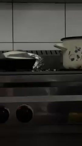 The camera slowly pans across a stainless steel stove top in a kitchen. There is a pan on one burner and a pot on another, with both partially covered by aluminum foil. White tiled walls are visible in the background.