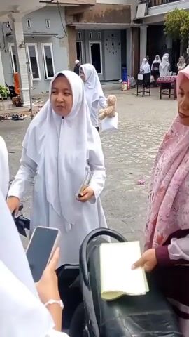 A young girl wearing a white hijab and tunic is standing and holding a dark-colored book or folder. She is facing a woman wearing a pink hijab and maroon top, who is holding a yellow piece of paper. The woman is looking at the girl. In the foreground, a hand holding a smartphone is visible, as if recording the interaction. A motorcycle seat is in the lower right corner of the frame. In the background, other people, some wearing hijabs, are seen in what appears to be an outdoor courtyard or common area, with buildings visible behind them. The ground is paved.