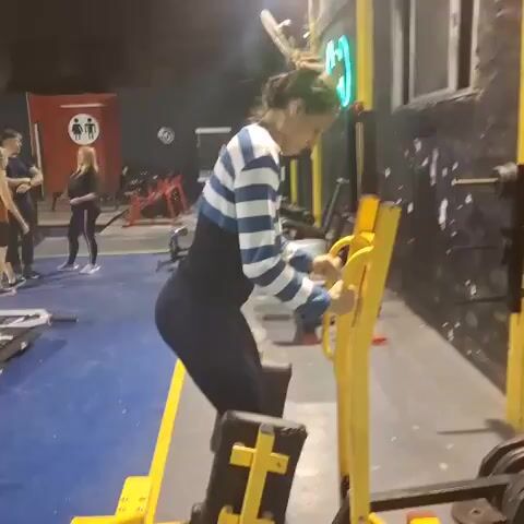 A woman is shown from a side and slightly rear angle performing a hip thrust or glute drive exercise on a yellow specialized machine in a gym setting. She wears a blue and white striped long-sleeved crop top over a black top, possibly a waist trainer, and black leggings. Her hair is tied up. She is actively pushing her hips upwards against the machine, which has weight plates loaded onto it, and then lowering them.

The gym features dark walls, a grey floor with a blue line, and mirrors in the background that reflect parts of the gym and the woman exercising. Another person, also in workout attire, is partially visible in the distant background. The lighting is artificial and typical of an indoor gym. No legible text is visible in the video. The activity takes place in a gym located in Wilde, Argentina.