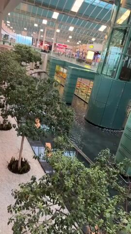 The video pans across the interior of a modern shopping mall. In the foreground, several potted trees with lush green foliage are visible, some featuring small, illuminated lights. The floor area to the left is covered with white gravel, and a tall, marble-clad pillar stands prominently.

The central feature of the mall appears to be a large, multi-level water feature. The water is dark and still, with some areas reflecting the lights above. A transparent glass elevator shaft rises from the water, encased in dark green panels.

Bridges or walkways with dark green structures span across the water. In the background, the upper levels of the mall are visible, featuring shop fronts with illuminated signs and displays. The ceiling is a high, translucent structure, allowing natural light to filter in, supplemented by numerous artificial lights. The overall impression is one of an open, airy, and contemporary shopping environment with integrated natural elements. There are no people clearly visible in the video.