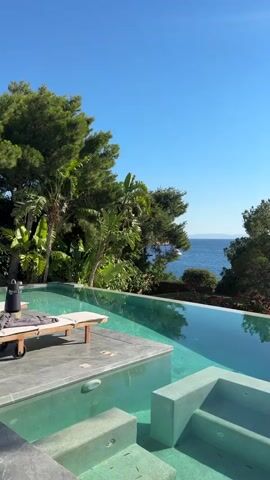 The video pans across a pristine infinity pool with clear turquoise water, overlooking a serene blue ocean under a clear sky. Lush green trees and foliage surround the pool, with a hint of distant land visible on the horizon. A modern chaise lounge sits on a concrete deck to the left of the pool. The water is still, reflecting the sky and surrounding greenery. The overall scene evokes a sense of tranquility and luxury.
