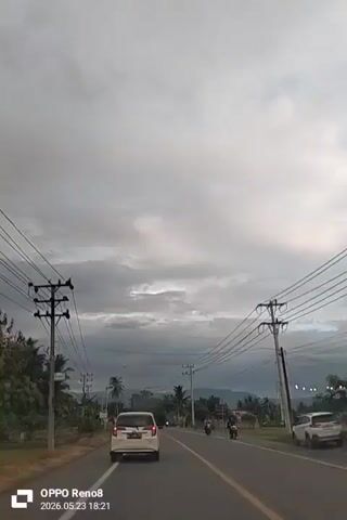 The video shows a view from inside a car driving on a two-lane road under an overcast sky. A white car is directly in front, moving in the same direction. Several motorcycles are also visible on the road. Power lines run alongside the road, and utility poles are sporadically placed. The surrounding environment includes trees, some of which appear to be palm trees. The road is marked with a double yellow line. A communication tower can be seen in the distance. The date and time "2026.05.23 18:21" are displayed at the bottom.