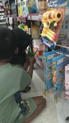 This media file shows two young children playing on the floor in what appears to be a store. The child on the right, wearing a green t-shirt and shorts, is partially visible from the side, facing left. The child on the left is wearing a black hijab and a green t-shirt. They are both looking at a small green alligator toy held by the child on the left.

To the left of the children, there are shelves stocked with various products. A prominent item is a yellow toy gun labeled "HOT SHOTS BANDIT" with red and yellow darts. Other items on the shelves are difficult to discern clearly but appear to be toys or packaged goods, some with colorful designs. Above the children, to the right, are more shelves with items in clear plastic packaging, featuring cartoonish characters and the word "MINI."

The floor is tiled with a light color. The lighting suggests an indoor environment, possibly a convenience store or a toy shop. The children appear engaged in play, with one child holding the toy and the other observing. There are no overt indications of time of day or weather. The overall scene is one of casual play within a retail setting.