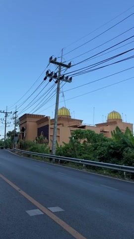 The video shows a building with elaborate architectural details. The building is mostly tan with some dark blue textured accents and features gold-colored domes on the roof. There is a large, ornate gateway leading into the property. The building is situated on a hillside surrounded by greenery, including banana trees and hedges. A black asphalt road runs in front of the building, with a yellow line marking the center. Power lines and utility poles are visible along the roadside, slightly obscuring the view of the building and sky. Several motorcycles are parked near the gateway. The sky is clear and blue.