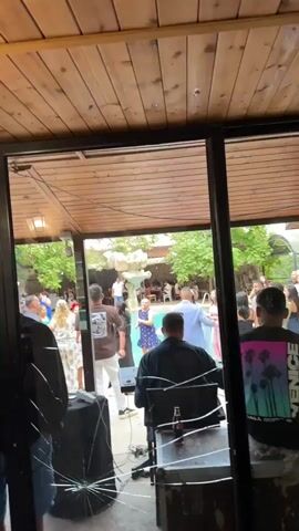 The video shows an outdoor party scene viewed through a large window. In the foreground, a person is partially visible from the back, seated at a table with a laptop and a red bottle. The background features a pool with a fountain and people dancing. A man is playing a clarinet, and another person is playing a keyboard. Guests are dressed in various casual and formal attire. Trees and an outdoor seating area are visible in the background. The ceiling above the window is made of wooden planks. The glass of the window has some cracks.