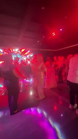 The video captures a lively party or event with people dancing in a dimly lit room. A large, bright LED screen displaying abstract colorful patterns serves as the backdrop. The lighting is dynamic, with purple and blue spotlights illuminating the dance floor and the guests. In the foreground, a person in a sparkling gold outfit is the central focus, seemingly dancing enthusiastically. Other individuals, some in formal attire like a white dress, are also visible on the dance floor, some dancing individually and others in pairs or groups. The floor is reflective, amplifying the colorful lights. The overall atmosphere appears festive and energetic.