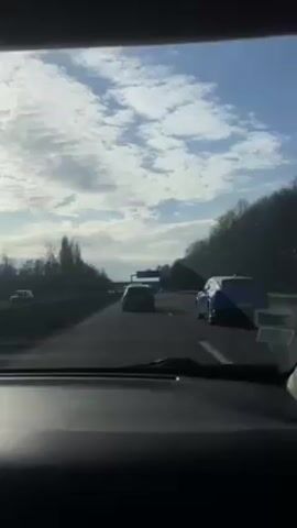 The video shows a car driving on a highway on a partly cloudy day.  Other cars are visible in the adjacent lanes. The highway is flanked by trees. A sign above the road is visible. The view is from inside a car.