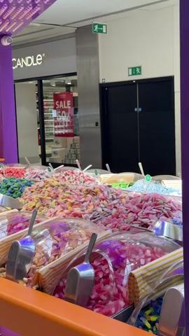 The video shows a close-up view of a large candy display in a store. The display is filled with a variety of colorful gummy candies, including pink, yellow, green, and blue varieties. Scoops are placed in each section of candy for customers to serve themselves. In the background, a store with the name "CANDLE" is visible, with a "SALE 50% OFF" sign in the window. A person in a black t-shirt and shorts walks past the candy display towards the store entrance. Another person is partially visible in the background. The overall environment appears to be an indoor shopping mall.