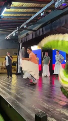 A group of people are on a stage. A man in an orange t-shirt and white pants is spinning in the center, holding a white fabric that expands around him. To his left, a man in a black and white shirt holds a microphone. To his right, two men and a woman are standing, all wearing white skirts. A woman in a green dress is in the foreground on the right, facing away from the camera. The stage has lights overhead and two screens with what appear to be flags displayed on them in the background. The floor of the stage is wooden and appears to be wet or reflective.