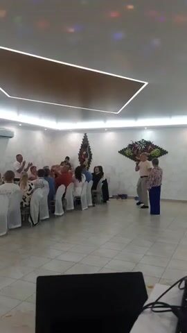 A group of people are gathered in a brightly lit room, possibly a banquet hall or event space. Most of the attendees are seated at long tables covered with white tablecloths, facing the camera. A man, standing and facing the seated guests, is speaking into a microphone. He is wearing a light-colored shirt and pants. To his right, a woman stands facing him and the guests, wearing a patterned blouse and blue pants. The room has white walls with a marble-like pattern and a modern ceiling with recessed lighting. Decorative floral arrangements are visible on the wall behind the speakers. Colorful lights are projected onto the ceiling. In the foreground, a portion of a black object, possibly a speaker or a table, is visible. The overall atmosphere suggests a social gathering or event.
