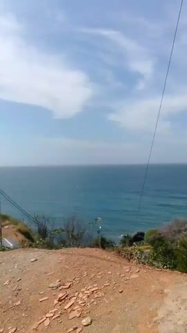 The video shows a wide shot of the ocean and sky. The camera is positioned on a dirt incline overlooking the water. In the foreground, there is dry dirt and some sparse vegetation. A thin black cable stretches diagonally across the frame from the upper right to the middle left. Below the cable and slightly to the right, there are a few buildings with white roofs. The ocean is a deep blue with gentle waves, and the sky is blue with scattered white clouds. The overall impression is a coastal landscape on a clear day.