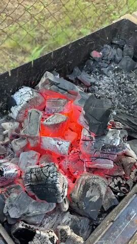 A metal barbecue grill with hot coals is seen in the video. The coals are glowing red in the center and covered in white ash on the outside. A red object is moved back and forth over the coals in a fanning motion, which causes sparks to fly. The grill is set on grass. A wire fence is visible in the background.