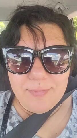 A selfie video shows a woman with dark hair wearing large black sunglasses and a gray and white patterned shirt. She is sitting in the passenger seat of a car, with a black seatbelt across her chest. The sun is shining, and the reflection of the outside scenery is visible in her sunglasses. The woman's mouth is slightly open, and she appears to be speaking. The interior of the car, including the ceiling and a portion of the door, is visible in the background. The video appears to be taken while the car is in motion, as evidenced by the slight movement and the blurred scenery in the reflections.