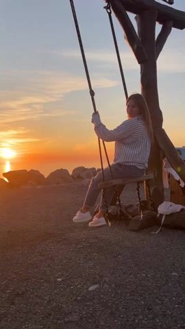 A young person swings on a rustic rope swing at sunset. The person, silhouetted by the bright sun and golden light over water, wears a striped shirt and jeans. The swing is attached to a wooden structure resembling a tree trunk. The ground is covered in small stones. A car is parked in the background.