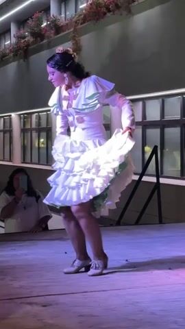 A young woman with dark hair pulled back and adorned with flowers is dancing flamenco on a wooden stage under purple and blue lights. She is wearing a white flamenco dress with ruffles and green trim, and white heeled shoes. Her right hand is holding the skirt of her dress, which flares out as she moves. Her left hand is raised to her face. In the background, a person wearing a white shirt is visible, holding a phone to take a picture. The environment appears to be outdoors at night, with a building with large windows and plants visible behind the stage.