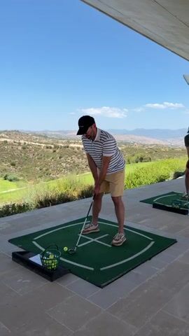 A man wearing a striped shirt, tan shorts, and a black baseball cap stands on a green golf mat with a golf club in his hands, preparing to swing. A basket of golf balls is to his left. He is on an outdoor patio with a view of rolling hills under a clear blue sky. Another person is partially visible to the right. The man speaks, stating "Wait guys, guys, don't shoot, don't shoot. Wait. Let me take video, okay? Wait, wait, I'm shooting."