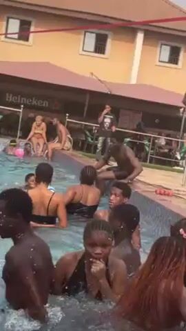 The video shows a group of people in and around an outdoor swimming pool. Many people are in the water, splashing and interacting. Some are wearing swimsuits. Others sit on the edge of the pool. The pool is surrounded by tile and what appears to be a building with a bar area. There is a sign with "Heineken" visible behind the pool. Some people are sitting at tables and chairs in the background. The weather appears to be sunny.